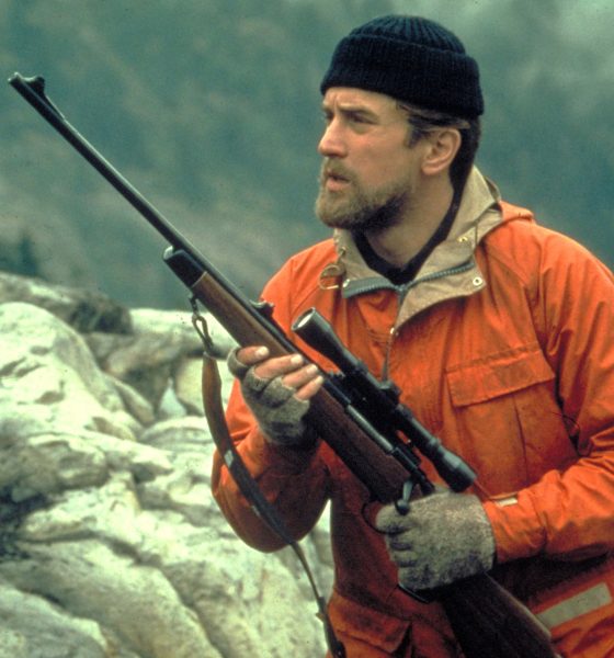 Robert De Niro in an orange jacket and deerstalker hat, carrying a scoped rifle in the film The Deer Hunter