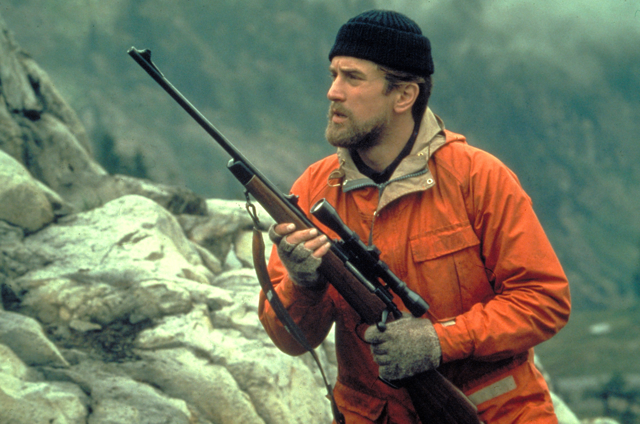 Robert De Niro in an orange jacket and deerstalker hat, carrying a scoped rifle in the film The Deer Hunter